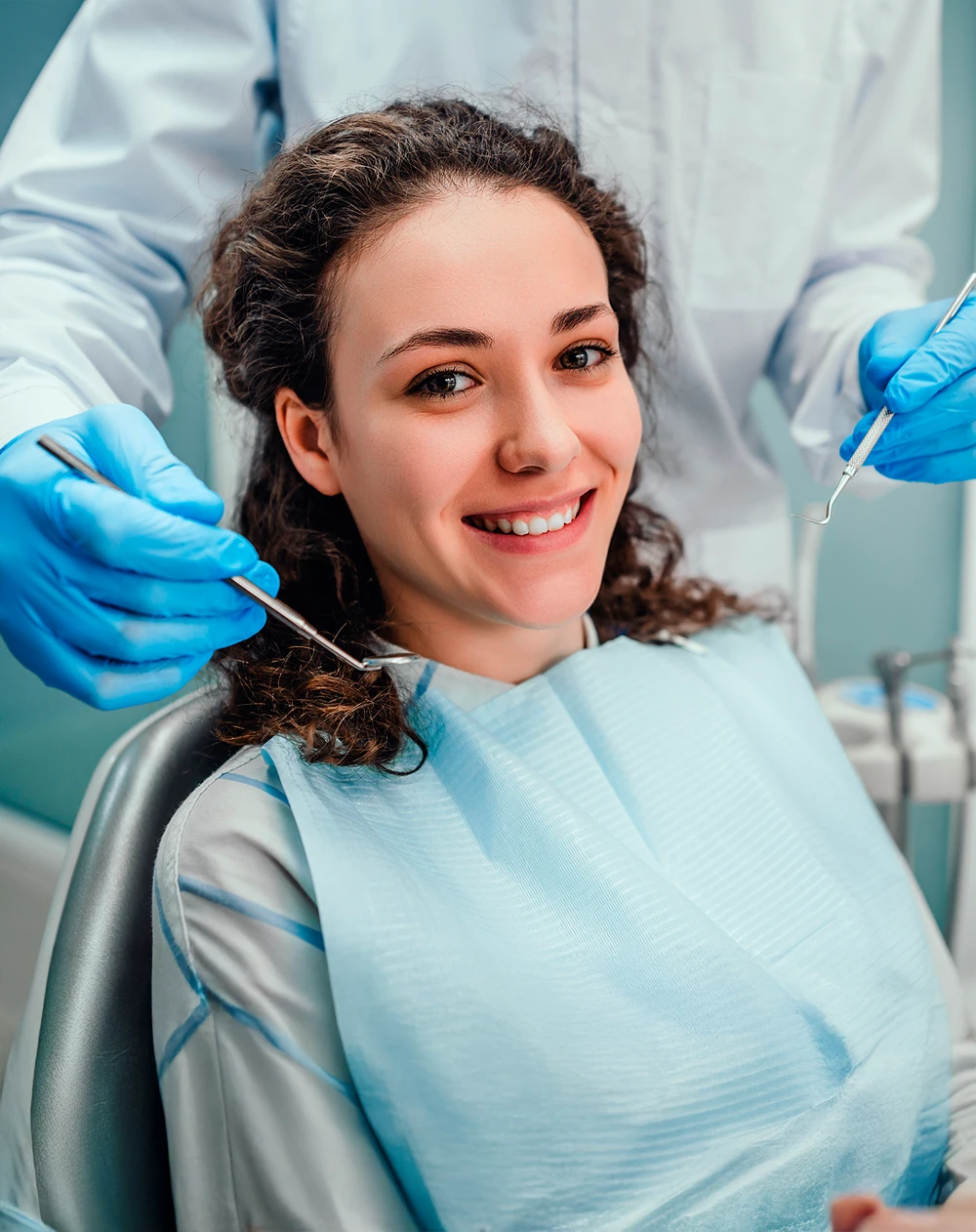 Person seated in a dental chair wearing a protective bib while gloved hands hold dental instruments nearby.